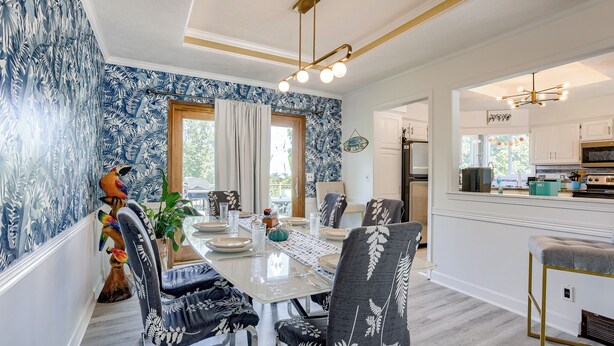 A dining area with a large table set for four, surrounded by chairs with a leaf pattern. There is a window with curtains, a plant on the table, and a kitchen visible in the background.