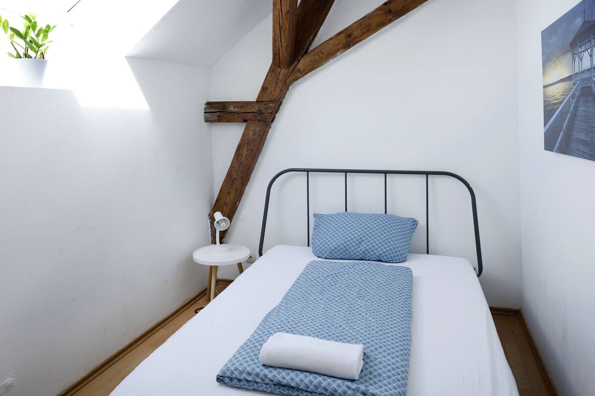 A small, neatly arranged room with a bed, a small table, and a potted plant.