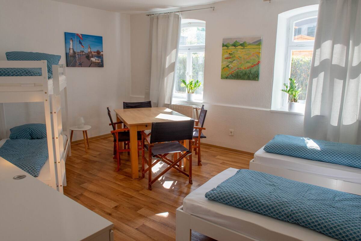 A room with bunk beds, a dining table, chairs, a window with curtains, and two potted plants.