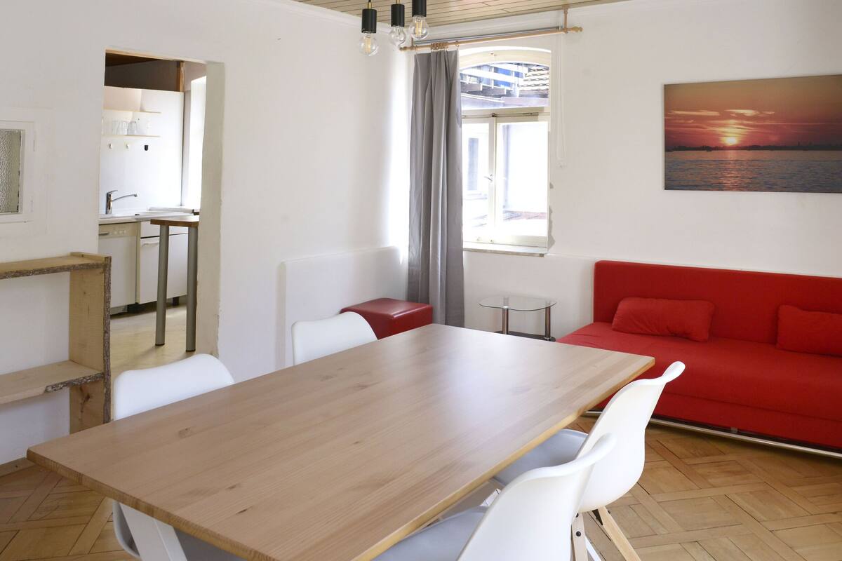 A room with a wooden table, white chairs, a red sofa, a small glass table, and a painting of a sunset.
