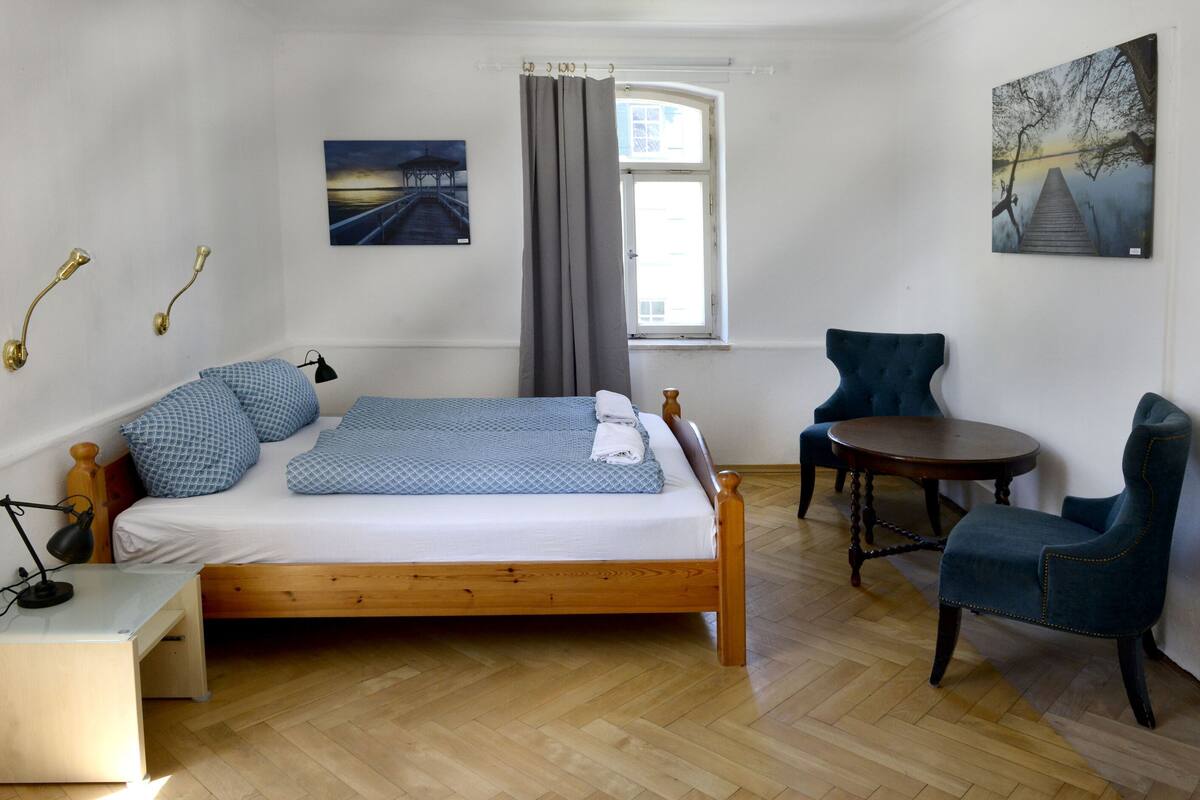 A bedroom with a wooden bed, a chair, a small table, and two paintings on the walls.