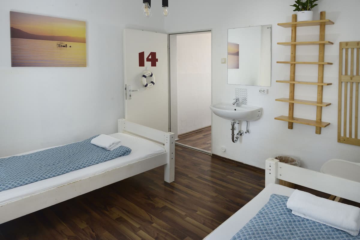 Une chambre avec deux lits simples, un lavabo blanc, une Ă©tagĂšre en bois en forme dâĂ©chelle, une porte portant le numĂ©ro 14 et un tableau reprĂ©sentant un coucher de soleil sur lâeau.