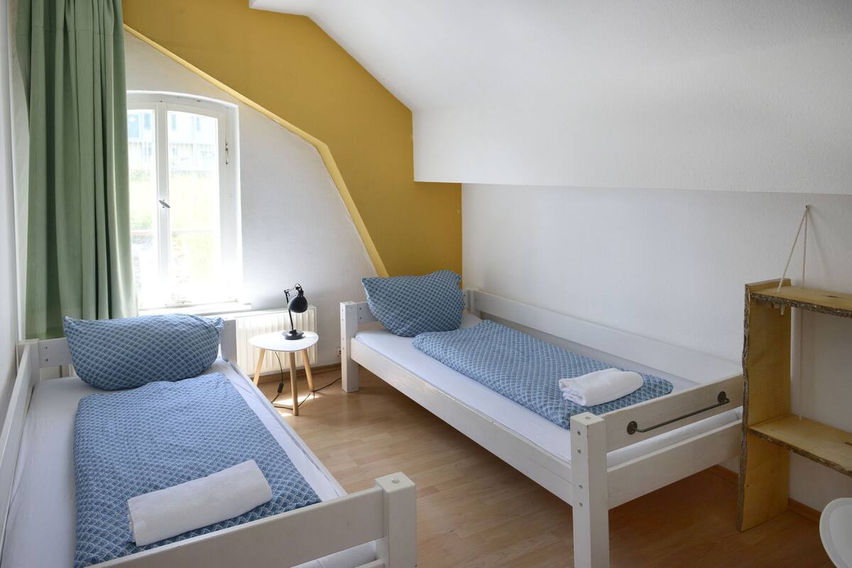 A dormitory room with two single beds, a small table with a lamp, a window with curtains, and a wooden shelf.