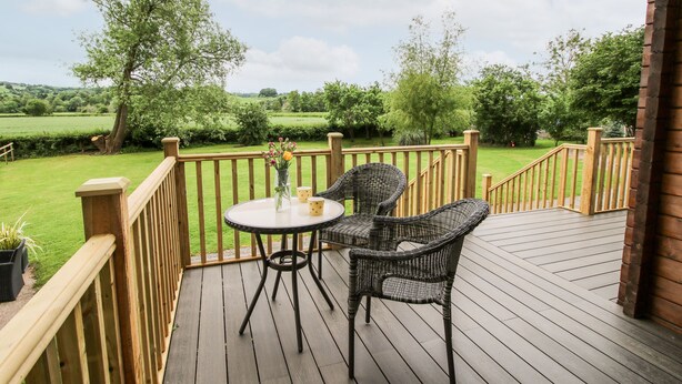 Um deck de madeira com uma mesa e duas cadeiras de vime, com vista para uma paisagem verde.