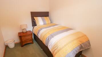 A small, neatly made bedroom with a single bed, a nightstand, a lamp, and a waste basket.