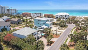 A coastal residential area with a mix of modern and traditional architecture, featuring a prominent blue building with a swimming pool, surrounded by palm trees and a clear view of the ocean.