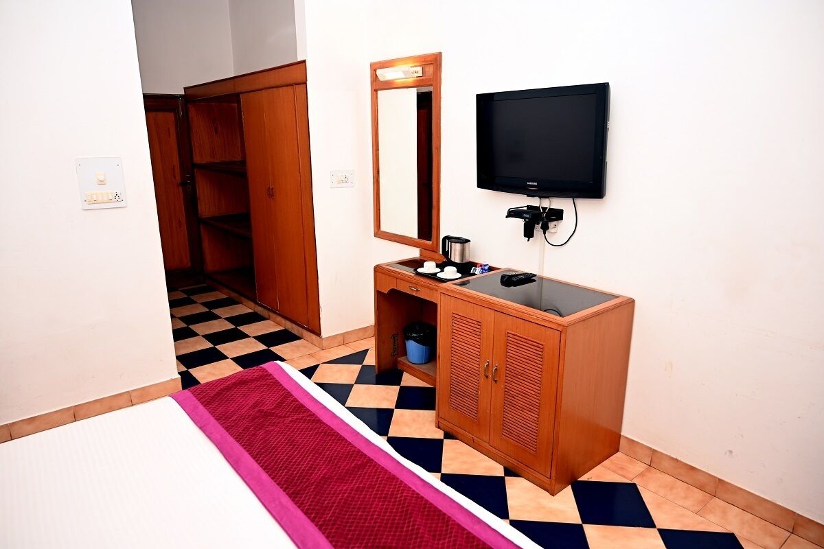 A hotel room with a checkered floor, a wooden cabinet with a mirror, a flat-screen TV, and a small table with a kettle and cups.