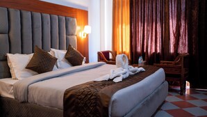 A hotel room with a large bed, a headboard, a nightstand, a lamp, a chair, and a window with curtains. - Hotel Hemkunth (Kasauli)