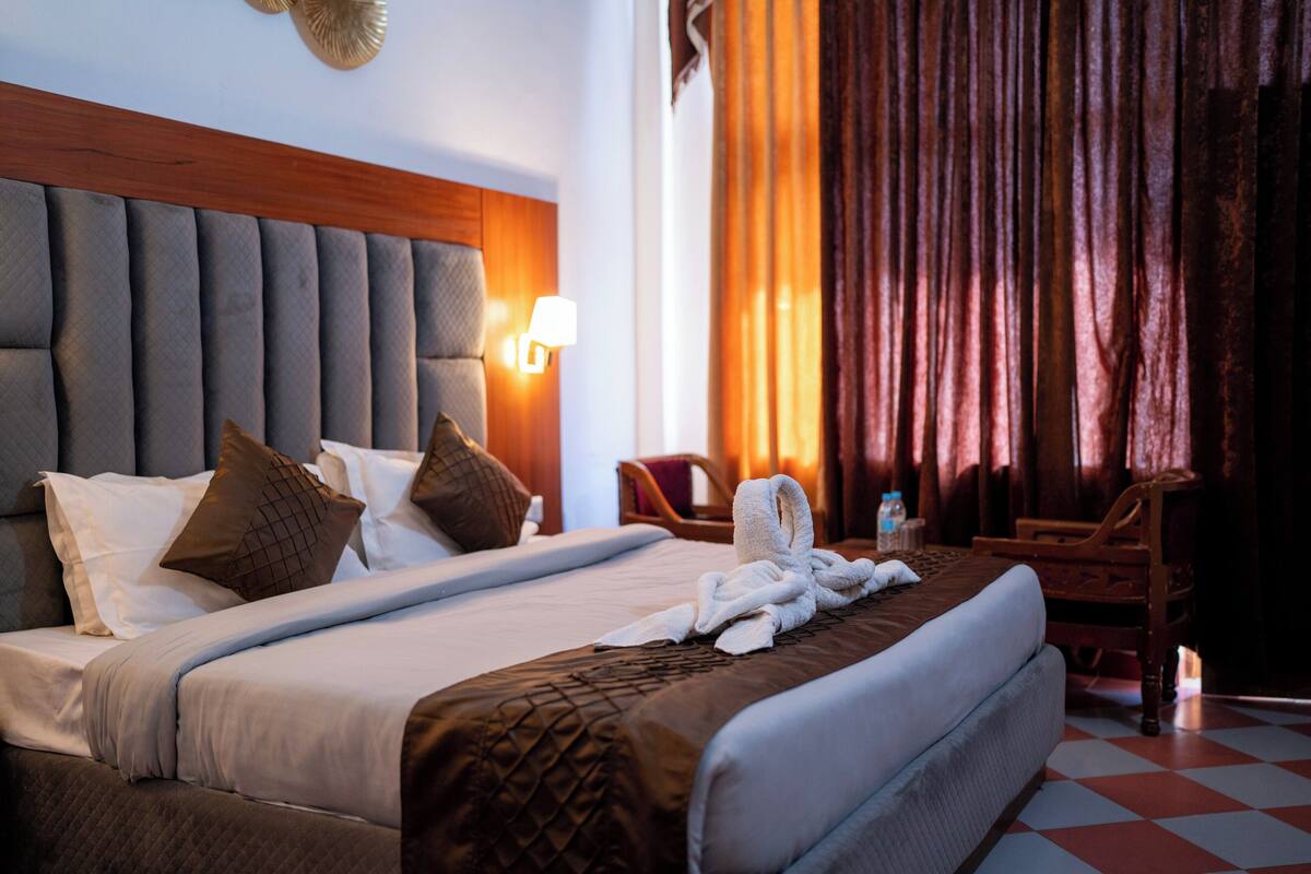 A hotel room with a large bed, a headboard, a nightstand, a lamp, a chair, and a window with curtains.