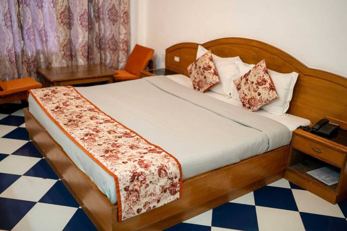 A hotel room with a checkered floor, a wooden bed with a floral bedspread, a nightstand with a phone, and a chair.
