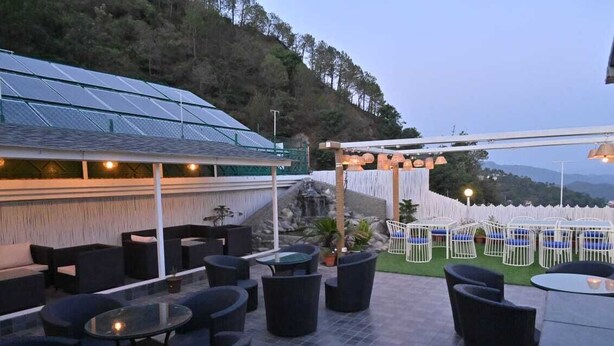 An outdoor patio area with black furniture, a pergola with hanging lights, and a view of a hillside. - Hotel Hemkunth (Kasauli)