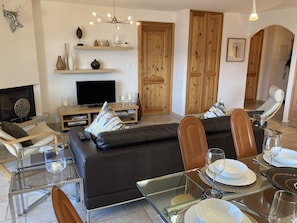 Una sala moderna con un sofá de cuero negro, una mesa de centro de vidrio y muebles de madera. Se observa un televisor, estanterías con objetos decorativos y una puerta de madera al fondo.