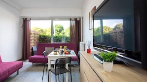 A modern living room with a sofa, dining table, chairs, a TV, and a large window with a view of greenery.