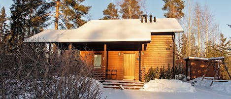 A wooden cabin with a covered porch, surrounded by snow and trees.