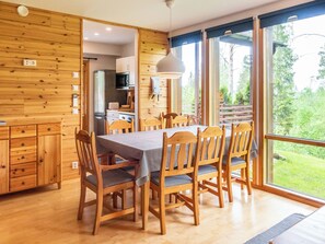 A wooden interior with a dining table and chairs, a kitchen area, and large windows with a view of greenery.