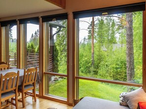 A room with a large window, a dining area with wooden furniture, and a view of a forest. - Kanervarinne by Interhome (Sotkamo)