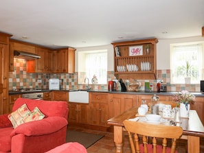 A kitchen with wooden cabinets, a red chair, a dining table with tea set, and a window with a plant. - Moonlight Cottage (Pwllheli)