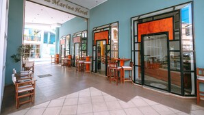 A hotel lobby with a large open space, multiple wooden tables and chairs, and glass doors leading to the outside. - Santa Marina Unique Hotel (Agios Nikolaos)