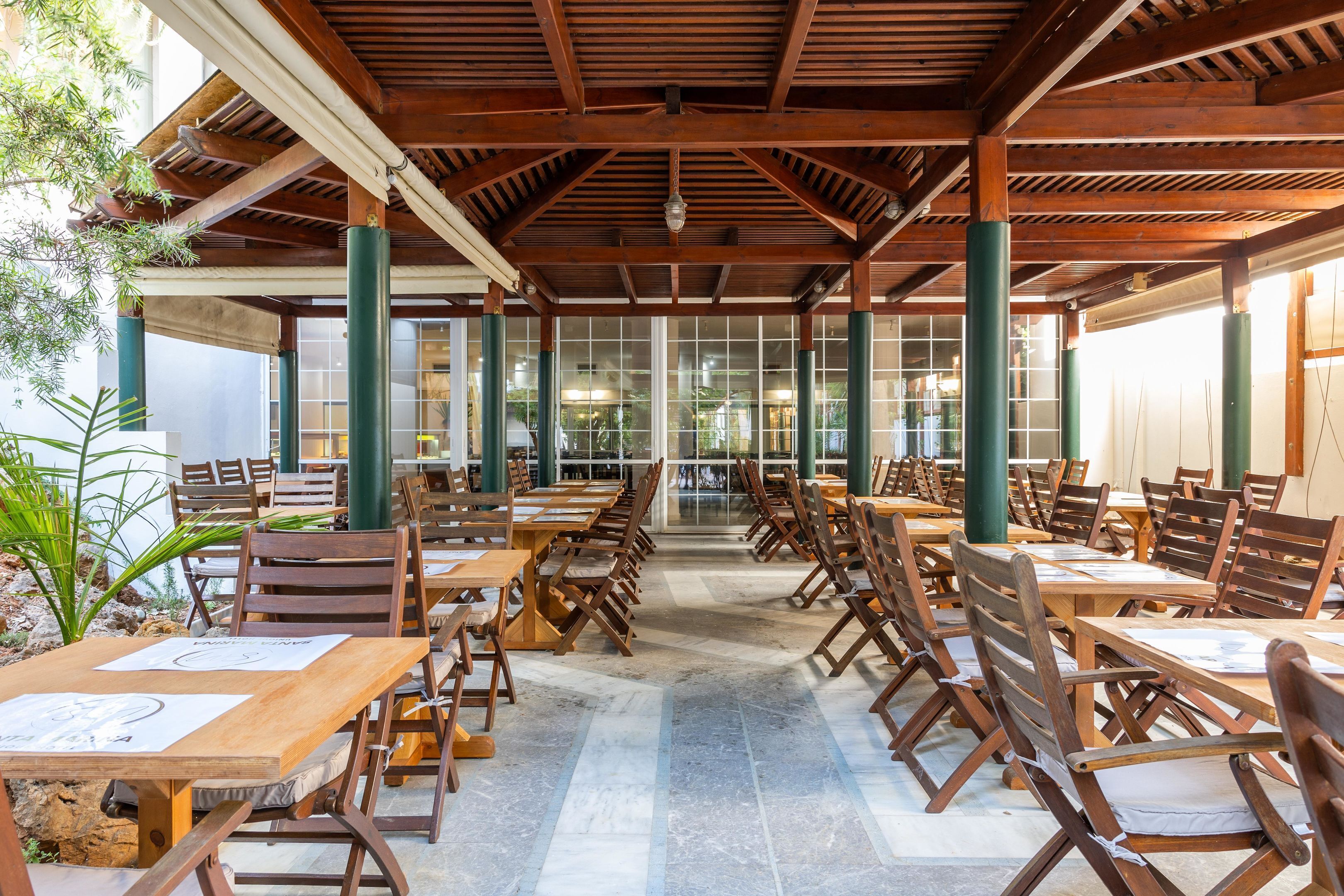 Un espacio de comedor al aire libre techado con mesas y sillas de madera, ventanas grandes y pilares verdes.
