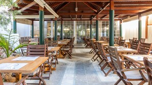 A covered outdoor dining area with wooden tables and chairs, large windows, and green pillars. - Santa Marina Unique Hotel (Agios Nikolaos)