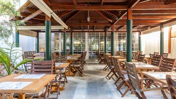 Un espacio de comedor al aire libre techado con mesas y sillas de madera, ventanas grandes y pilares verdes.