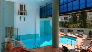 An indoor swimming pool with a view of a cityscape, a staircase leading to a balcony, and a large vase on the poolside. - Santa Marina Unique Hotel (Agios Nikolaos)