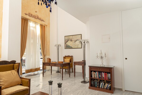 A living room with a sofa, a coffee table, a dining table with chairs, a bookshelf, a painting on the wall, and a door. - Santa Marina Unique Hotel (Agios Nikolaos)
