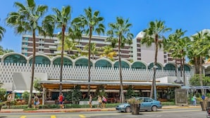 Een hotel met een moderne gevel, palmbomen en een overdekt terras.