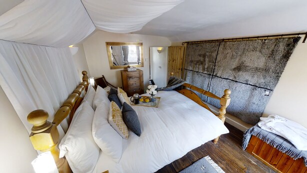 A bedroom with a wooden bed, white linens, a bedside table with a lamp, a dresser, and a stone wall.