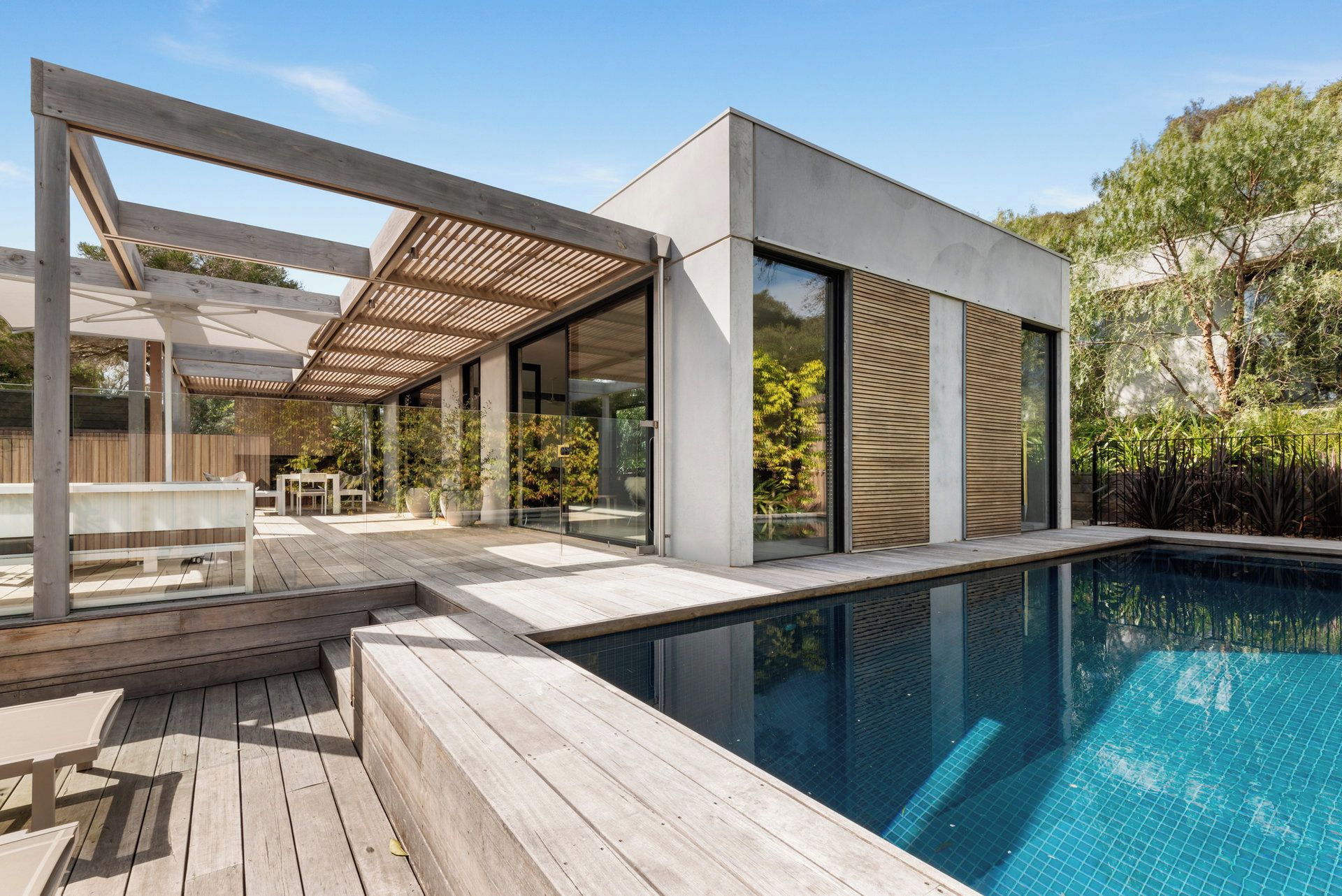 A modern outdoor pool area with a wooden deck, seating, and a glass-enclosed structure.