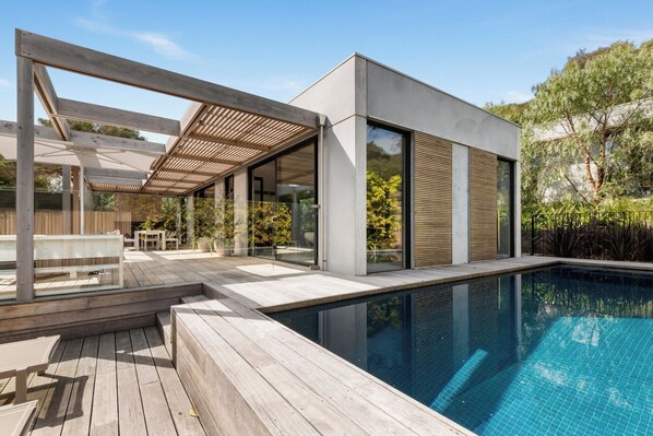 A modern outdoor pool area with a wooden deck, seating, and a glass-enclosed structure.