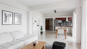 A modern living room with a white sofa, a wooden coffee table, and a black ottoman. There is a dining area with a white table and chairs, and a kitchen with red cabinets and a white countertop. - Alluring 2BR Apartment in Marousi (Marousi)