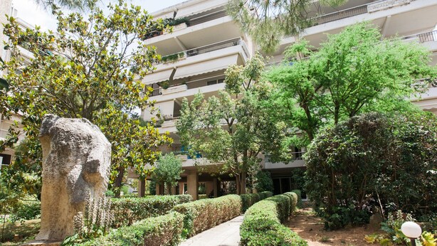 A courtyard with a stone sculpture, neatly trimmed hedges, and a multi-story building with balconies. - Alluring 2BR Apartment in Marousi (Marousi)