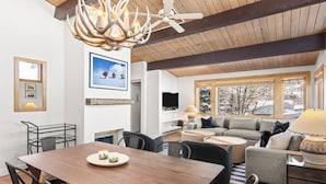 Een ruime woonkamer met houten plafond, een eettafel met stoelen, een bank en een televisie.