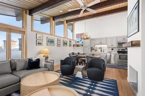 A modern living room with a grey sofa, two armchairs, a wooden coffee table, and a dining area with a table and chairs. A kitchen with white cabinets and stainless steel appliances is visible in the background.