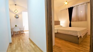 A hotel room with a wooden floor, a bed with white bedding, a small table with a chair, and a wall clock. - Apartment IPEK (Budva)