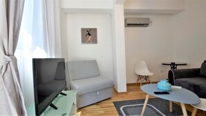 A modern living room with a grey sofa, a small round table, a TV on a glass stand, a white chair, and an air conditioning unit on the wall. - Apartment IPEK (Budva)