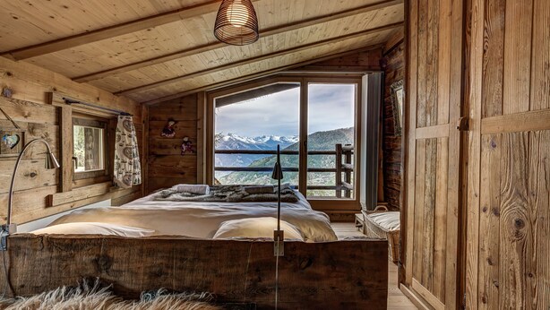 Un dormitorio de cabaña de madera con una cama grande, una ventana con vistas a la montaña y una lámpara colgante.