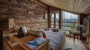 Un dormitorio de cabaña de madera con una cama, una silla, un escritorio con un libro y una ventana con vistas a la montaña.