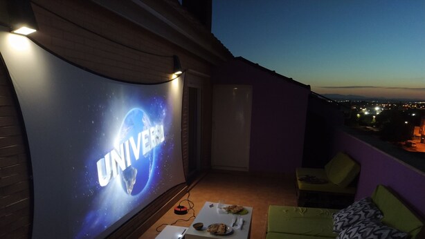 Un balcón con una pantalla de proyector mostrando la palabra "UNIVERSAL", una mesa con un plato y una copa, y un sofá verde con cojines estampados.