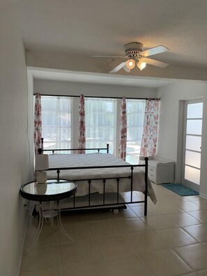 Ein Schlafzimmer mit einem Bett, einem Deckenventilator, einem Fenster mit Vorhängen, einem kleinen Tisch und einem Stuhl.