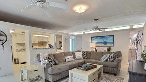 Un salon comprenant un canapé d’angle, une table basse et une table d’appoint. On y trouve un ventilateur de plafond, un tableau accroché au mur et, par une porte ouverte, on peut apercevoir la cuisine.
