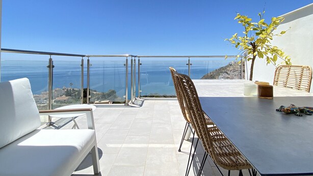 Un balcón con vista al mar, una mesa con sillas y un pequeño árbol.