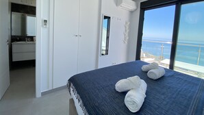 Un dormitorio con una cama grande, vista al mar, un armario blanco y un baño con espejo y lavamanos.