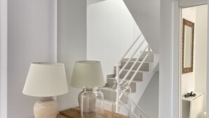 Un interior moderno con una mesita de madera que sostiene dos lámparas y un florero de vidrio. Se observa una escalera con un diseño geométrico en la pared de fondo.