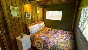A room with wooden walls, a bed with a colorful patterned bedspread, a small bedside table with a plant, and framed pictures on the wall. - Awe Inspiring two Storey Tent (Battle)
