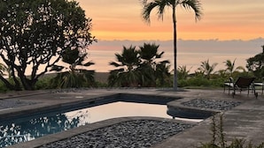 A pool with a curved edge, surrounded by pebbles and plants, with a view of the sunset over the water. - Kohala Estate Home 3 Bedroom Estate by RedAwning (Kamuela)