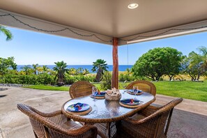 An outdoor dining area with a view of the ocean, wicker chairs, and a table set with plates and glasses. - Kohala Estate Home 3 Bedroom Estate by RedAwning (Kamuela)