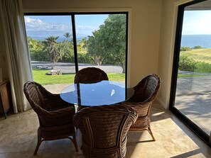 Una habitación con una mesa de vidrio y sillas de mimbre, un ventanal con vistas al océano y al follaje.