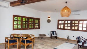 Una habitación con una mesa de comedor de madera y sillas, una mesa redonda de centro, un tapete estampado y ventanas grandes con marcos de madera.